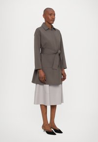 Frau in einem taubfarbenen, mit Gürtel versehenen Mantel über einem hellgrauen Midirock und schwarzen spitzen Slingback-Schuhen, stehend vor einem schlichten weißen Hintergrund.