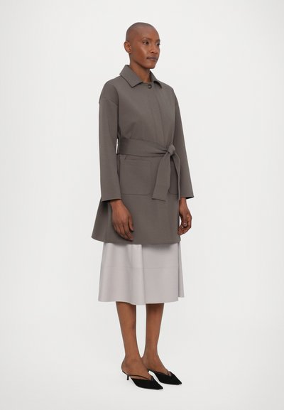 Frau in einem taubfarbenen, mit Gürtel versehenen Mantel über einem hellgrauen Midirock und schwarzen spitzen Slingback-Schuhen, stehend vor einem schlichten weißen Hintergrund.