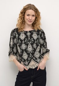 Svart blus med intrikata vita blommönster och paisleymönster, med spetsdetaljer på ärmar och nederkant. Tyget verkar lättviktigt.