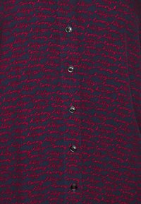 Navy cardigan featuring red embroidered text of "Tommy Hilfiger." Six black buttons down the front, textured fabric with a repeating pattern.