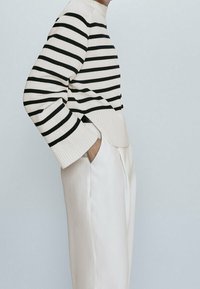 Pull en maille rayé crème et noir, doté d'un col haut, de poignets côtelés et d'ouvertures sur les côtés, associé à un pantalon tailleur de couleur claire.