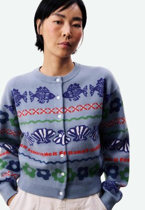 Vrouw die een lichtblauw vest draagt met patronen van donkerblauwe vissen, schelpen, groene en blauwe bloemen, en rode decoratieve lijnen, handen in de zakken.