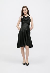 Μαύρο φόρεμα με λαιμόκοψη καπό από λείο, γυαλιστερό ύφασμα. Εφαρμοστό μπούστο με φουσκωτή φούστα, ασύμμετρο τελείωμα και λεπτές τιράντες.