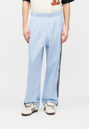 Άτομο που φορά ανοιχτό μπλε παντελόνια φόρμας με μαύρες πλευρικές ρίγες και λευκά αθλητικά παπούτσια με μαύρες λεπτομέρειες σε μονόχρωμο φόντο.