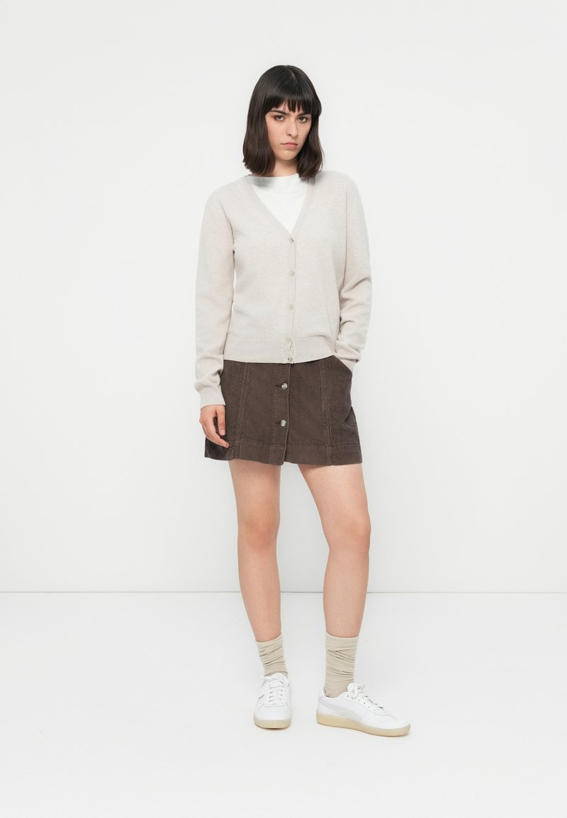 Donna con cardigan beige sopra una maglietta bianca, gonna marrone con bottoni, calze beige e sneakers bianche, in piedi con una mano in tasca.