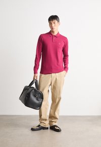 Bordeaux langwerpige polo met een zak, beige broek, zwarte leren weekendtas en zwarte schoenen met uitsnijdingen. Eenvoudige moderne stijl.