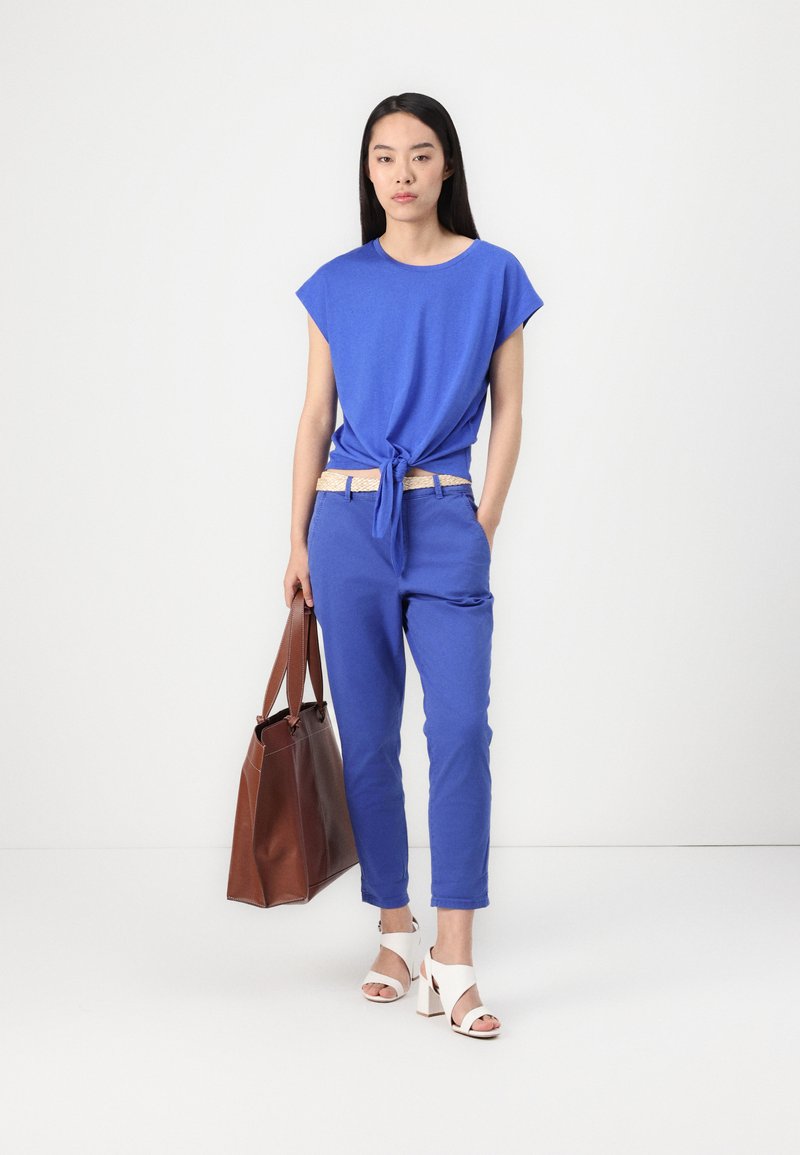 Blaue T-Shirt mit Knoten vorne, passende blaue Hose, braune Ledertasche und weiße Sandalen mit Blockabsatz. Glattes Material, lockere Passform.