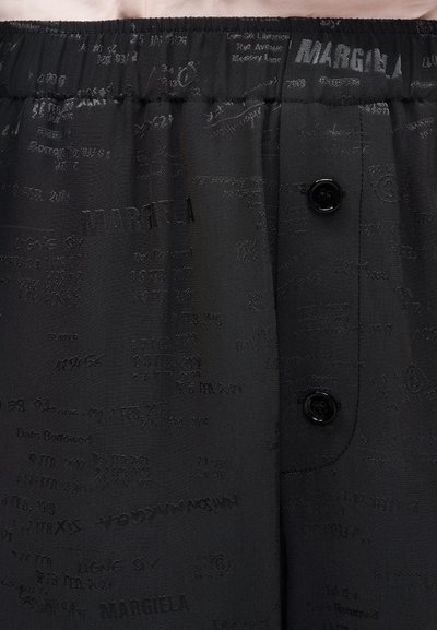 Κοντινό πλάνο μαύρου υφάσματος με λεπτό επαναλαμβανόμενο κείμενο "MARGIELA" και μικρά μαύρα κουμπιά στη μανσέτα με ρυτίδες.