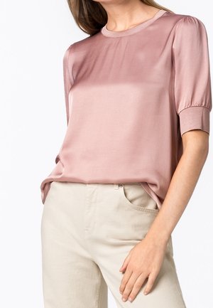 Femme portant un chemisier en satin rose poudré à manches courtes, rentré dans un pantalon crème taille haute, main posée sur la hanche.
