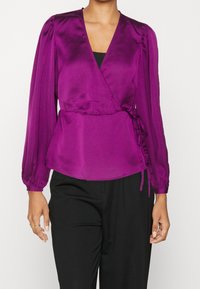 Blusa avvolgente in satin viola con maniche lunghe a palloncino, scollo a V e dettaglio con nodo laterale; texture liscia, aderente in vita, indossata sopra pantaloni neri.