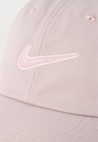 Ροζ ανοιχτό καπέλο baseball με κεντημένο λογότυπο Nike swoosh στο μπροστινό μέρος, που δείχνει την υφή του υφάσματος και τις λεπτομέρειες της ραφής.
