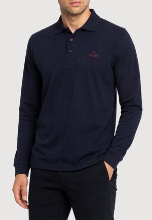 Mann trägt ein dunkelblaues langärmeliges Poloshirt mit drei Knöpfen und rotem "GISADA" Logo auf der linken Brust, kombiniert mit schwarzer Hose.