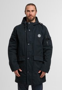 Black hooded parka made of sturdy fabric. Features large front pockets, button closure, drawstring hood, and a zippered pocket on the sleeve.