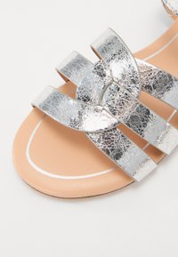 Silver sprucken konstläder sandal med tre breda remmar och en persikofärgad sula, med vit kontrastsöm och platt design.