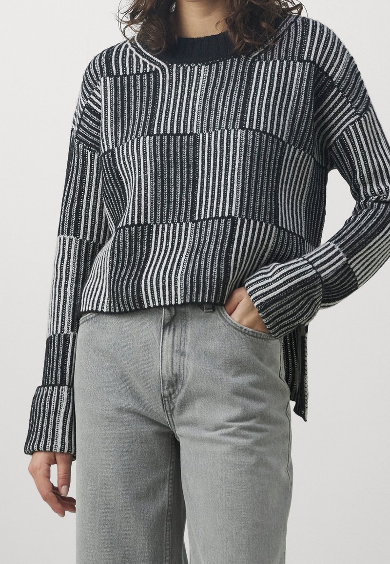 Maglione a maglia con righe bianche e nere; presenta una vestibilità comoda, spalle scese e polsini a costine. Indossato con jeans grigio chiaro.