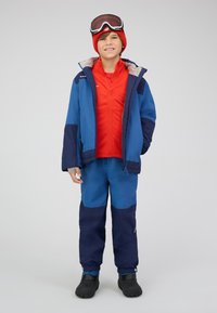 Ragazzo in piedi con giacca e pantaloni invernali blu e navy, maglione rosso con zip, berretto rosso, occhiali da sci sulla fronte e stivali da neve neri.