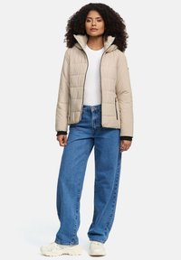 Vrouw met krullend haar in een beige pufferjack, een witte blouse, een spijkerbroek met wijde pijpen en witte sneakers, staande tegen een effen achtergrond.