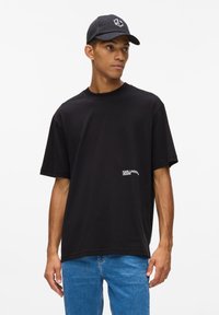 Melna kokvilnas T-krekls ar brīvu piegriezumu, ar mazu baltu logotipu tuvu apakšmalai. Kombinēts ar gaiši ziliem džinsu biksēm un melnu cepuri.