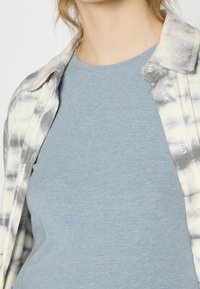 Ljusblå bomullströja under en skjorta med knappar med ett grått och vitt tie-dye-mönster, med stora knappar och vikbar krage.