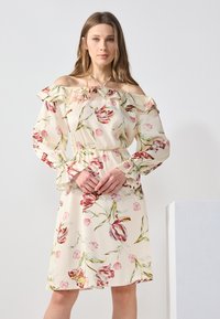 Robe dénudée sur les épaules en tissu léger crème avec un imprimé floral dans des teintes de rose et de vert. Présente un décolleté et des manches volants.