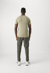 Hombre con camisa de manga corta beige claro y pantalones verde oscuro de pie, de espaldas, frente a un fondo claro. Zapatillas blancas visibles.