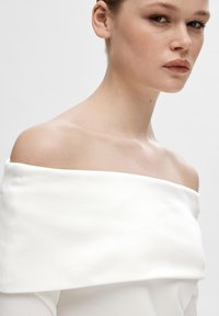 Hvit av-shoulder topp med en myk, jevn tekstur. Har en bred utringning og tettsittende design som fremhever kragebeinet.