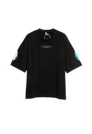 Joven con una camiseta negra holgada con estampados en las mangas, pantalones cortos blancos con detalles de estrellas negras, calcetines blancos y zapatos beige sin cordones con textura.