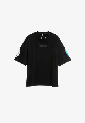 Joven con una camiseta negra holgada con estampados en las mangas, pantalones cortos blancos con detalles de estrellas negras, calcetines blancos y zapatos beige sin cordones con textura.