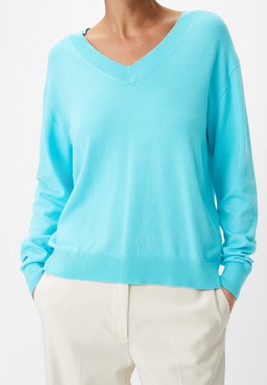 Žena oblečená v světle modrém svetru s výstřihem do V a dlouhými rukávy a krémových kalhotách, ruce v kapsách, na bílém pozadí.