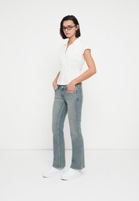 Λευκό πουκάμισο με κοντά μανίκια και κάθετες πιέτες στο μπροστά μέρος, συνδυασμένο με ανοιχτό μπλε ξεχειλωμένο τζιν και λευκά αθλητικά παπούτσια. Απλό, καθημερινό στυλ.