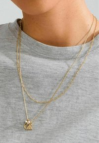 Gelaagde gouden kettingen met een gestructureerde schakel en een driehoekige hanger met parelaccenten, wat zich afspeelt tegen een grijs shirt.