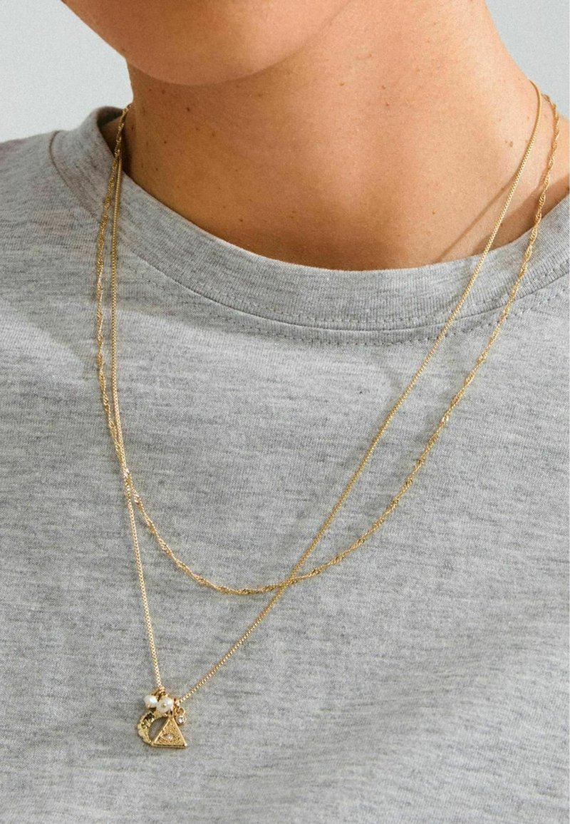 Gelaagde gouden kettingen met een gestructureerde schakel en een driehoekige hanger met parelaccenten, wat zich afspeelt tegen een grijs shirt.