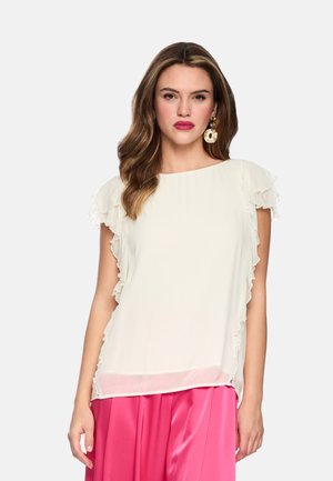 Woman with wavy hair wears white ruffled cap sleeve blouse and bright pink satin skirt, accessorized with large gold statement earrings.