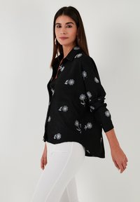 Black button-up shirt with white floral patterns, long sleeves, slightly loose fit, made of a lightweight fabric, worn with white pants.