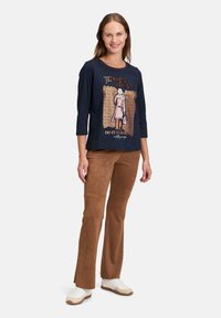 Navyblaues Langarmshirt mit Grafikdruck, kombiniert mit braunen Wildleder-Bootcut-Hosen und weißen Sneakers mit beigen Akzenten.