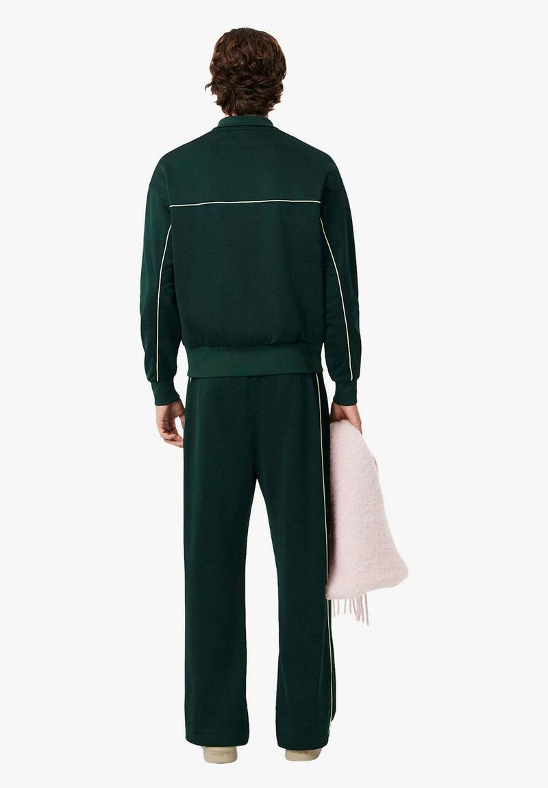 Tuta sportiva verde con piping bianco, composta da giacca con zip e pantaloni ampi. La persona tiene in mano una coperta soffice rosa chiaro.