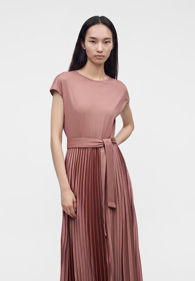 Femme aux longs cheveux foncés porte une robe plissée rose poudré avec une ceinture nouée à la taille, debout devant un fond clair uni.