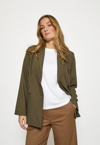 Olivovo zelené sako s prispôsobeným strihom, zapínaním na jeden gombík a zárezovými klopami, v kombinácii s bielou košeľou a hnedými nohavicami.