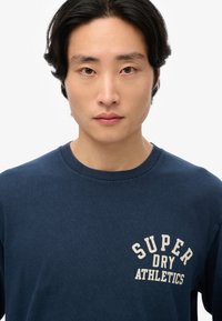 Camiseta de algodón azul marino con cuello redondo, que presenta el texto en color crema "SUPER DRY ATHLETICS" en la parte izquierda del pecho.