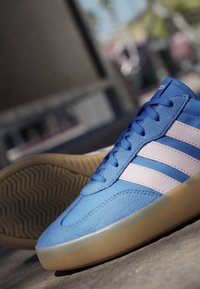 Blue Adidas sneaker with white stripes and gum sole placed on ground, urban background blurred.