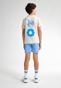 Camiseta de algodón beige con gráficos de llamas azules y engranajes en la parte posterior, combinada con pantalones cortos azul claros y zapatillas blancas con acentos verdes.