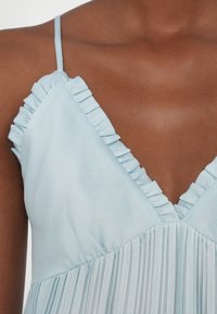 Jasnoniebieska sukienka z dekoltem w serek, ozdobiona falbanami wzdłuż linii szyi oraz plisowaną fakturą na górze. Regulowane ramiączka.