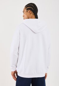 Hvit hettegenser med kengurulomme og justerbar snorhette, med ribbestrikkede mansjetter og nederkant, brukt med blå denim.