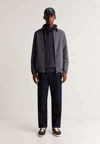 Veste légère grise sur un pull en tricot texturé marine, assortie à un jean en denim foncé et des sneakers grises avec des semelles blanches.