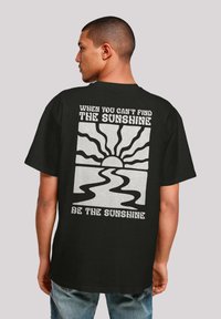 Schwarzes Baumwoll-T-Shirt mit einem grafischen Druck auf der Rückseite, der eine Sonne und ein gewelltes Flussdesign zeigt, begleitet von weißem Text.