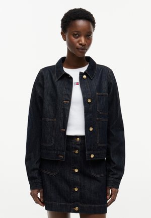 Kvinde stående iført mørk denimjakke og matchende knappet denimskørt over hvid skjorte med lille logo på brystet.