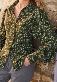 Donna che indossa una camicia verde a fiori con maniche a sbuffo e pantaloni grigi, in piedi contro un muro di pietra strutturato.