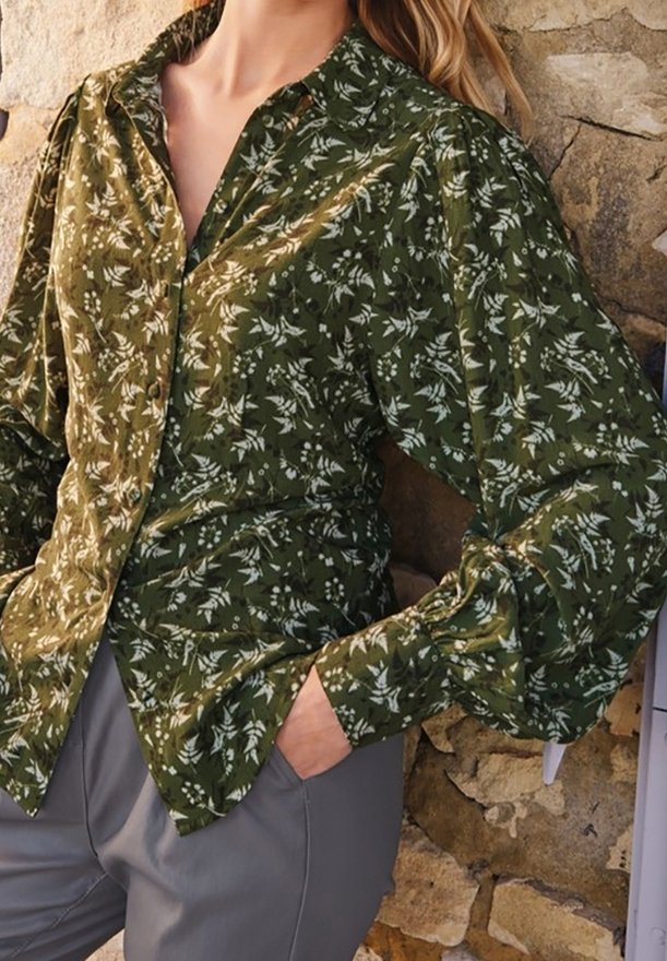Donna che indossa una camicia verde a fiori con maniche a sbuffo e pantaloni grigi, in piedi contro un muro di pietra strutturato.