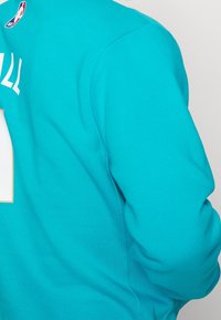 Felpa teal con collo rotondo, maniche lunghe e un grande numero bianco sul retro. Etichetta del logo NBA visibile nell'area del colletto.