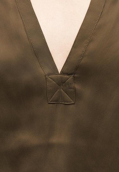 Μπλούζα από ελιά πράσινο ύφασμα με βαθύ V λαιμόκοψη και ενισχυμένη ορθογώνια ραφή στη βάση του λαιμού, σε ανοιχτόχρωμο δέρμα.